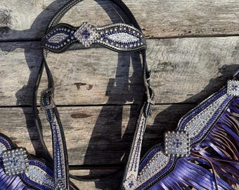 May include: A black leather horse bridle and breast collar set featuring purple and clear crystal embellishments. The browband and breast collar are adorned with silver-toned metal hardware, square crystal accents, and long purple leather fringe.