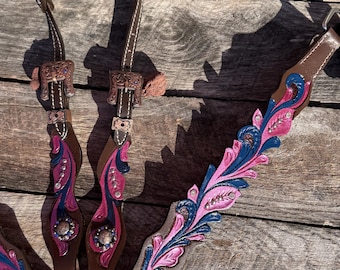 Puede incluir: Un cabezal de caballo de cuero marrón con intrincados grabados florales y detalles pintados a mano en rosa y azul. La pieza incluye hebillas de metal plateado, conchos decorativos y pequeños cristales incrustados en los diseños.