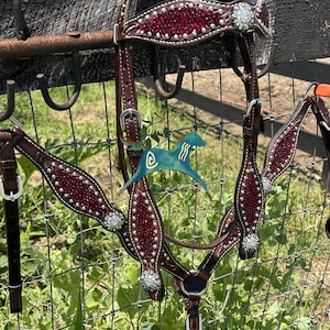 May include: A dark brown leather horse bridle and breast collar set featuring deep red crystal inlays and clear rhinestone accents. The tack includes silver-toned buckles and hardware, with a decorative browband and chest piece design.