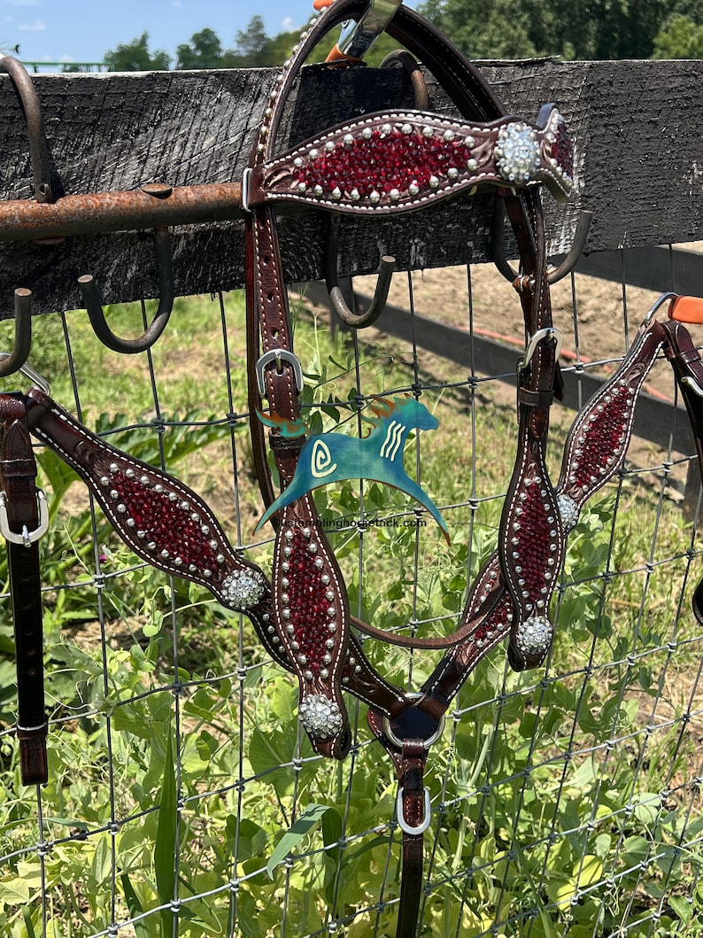 May include: A dark brown leather horse bridle and breast collar set featuring deep red crystal inlays and clear rhinestone accents. The tack includes silver-toned buckles and hardware, with a decorative browband and chest piece design.
