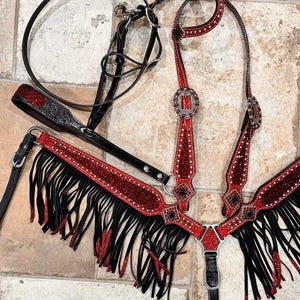 May include: A horse tack set featuring a black leather bridle and a red and black breast collar. The breast collar has black fringe and red glitter accents. The bridle and breast collar are adorned with silver-toned hardware and rhinestones.