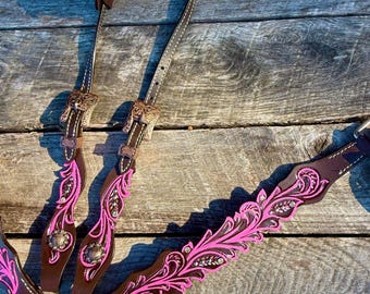 Puede incluir: Una brida de caballo de cuero marrón con bordado floral rosa y herrajes plateados. La brida presenta costuras decorativas y correas ajustables. El diseño incluye un peto y un cabezal.
