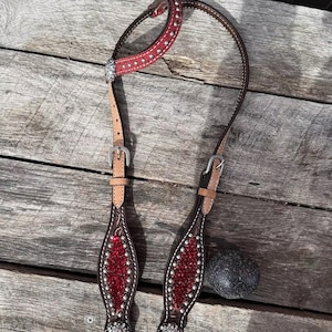 May include: A Western-style horse headstall with a red and brown leather design. The headstall features silver-colored studs and decorative accents. The leather is stitched with white thread, and the headstall has a buckle closure.