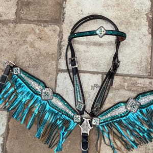 May include: A dark brown leather horse bridle and breast collar set featuring bright turquoise fringe, silver-toned hardware, and decorative square accents embedded with clear crystals. The set includes a browband with a central crystal detail.
