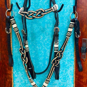 Puede incluir: Un juego de brida de caballo en blanco y negro con detalles dorados. La brida presenta intrincados diseños de nudos y costuras decorativas. El conjunto incluye un cabezal, una testera y un peto, todo colgado sobre un fondo turquesa.