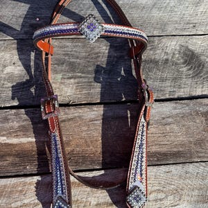 Könnte beinhalten: Ein brauner Lederpferdezaum mit silbernen Schnallen und dekorativen Strasssteinen. Stirnband und Backenstücke sind mit blauen, weißen und lila Strasssteinen verziert. Ein silbernes rautenförmiges Ornament befindet sich in der Mitte des Stirnbandes.