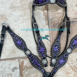 Puede incluir: Un conjunto de brida y petral para caballo en cuero negro con grabados florales morados, tachuelas plateadas y cristales transparentes. La testera y el petral presentan patrones de flores detallados y el texto customblinghorsetack.com.