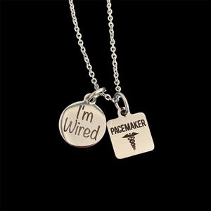 May include: A silver necklace with two pendants. One pendant is a round charm with the words "I'm Wired" engraved on it. The other pendant is a square charm with the word "Pacemaker" and a medical symbol engraved on it.