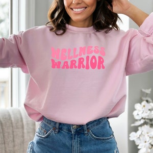 May include: Pink sweatshirt with the text "WELLNESS WARRIOR" in pink, retro-style lettering.