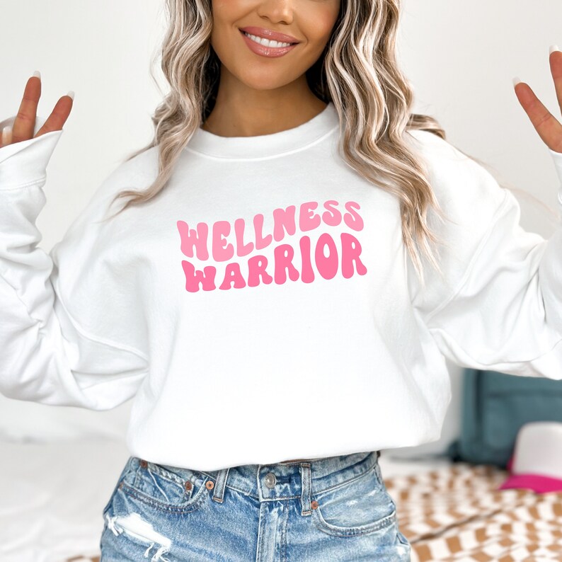 May include: White sweatshirt with a pink graphic that says "WELLNESS WARRIOR" in a retro font.