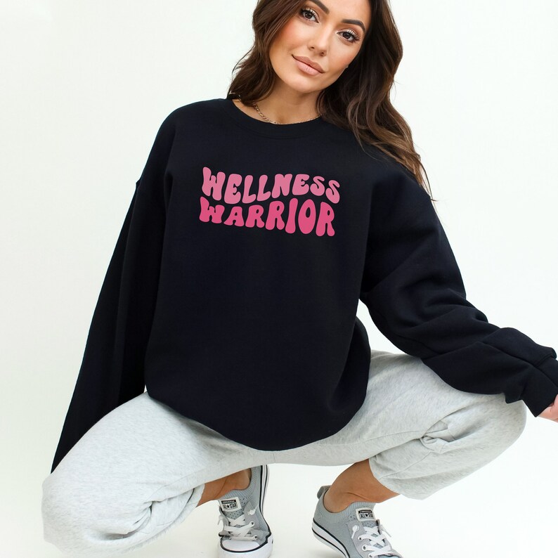 May include: A black sweatshirt with a pink "Wellness Warrior" graphic in a retro font.