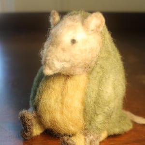 May include: A handmade felted animal figurine, featuring a light beige head, a yellow belly, and a green back. The small creature has tiny dark brown feet and ears. The toy is sitting on a wooden surface.