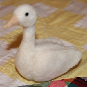 May include: A white felt swan with a small black eye. The swan is sitting on a yellow and white patterned fabric.