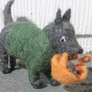 May include: A needle-felted gray dog figurine wearing a green sweater. The dog has pointed ears, a dark nose, and white-and-black eyes. It is holding a small, round, orange felted basket in its mouth, which contains a tiny white and brown object.