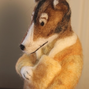 May include: A handmade felted dog figurine with a brown and white head, wearing a yellow coat. The dog's arms are crossed, and it has a serious expression. The figurine is a unique piece of art.
