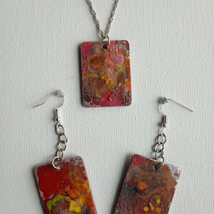 Può includere: Un set di gioielli composto da una collana e orecchini con ciondoli rettangolari. I ciondoli sono dipinti con motivi astratti in tonalità di rosso, arancione, giallo, marrone e bianco.