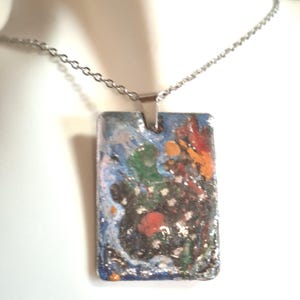 Può includere: Una collana con pendente rettangolare e catena argentata. Il pendente presenta un design astratto colorato con accenti blu, arancioni, verdi e bianchi. Il pendente misura circa 3,8 cm di lunghezza e 2,5 cm di larghezza.