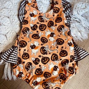May include: A peach-colored baby onesie with a Halloween-themed print featuring Winnie the Pooh, Tigger, and pumpkins. The onesie has black and white gingham ruffle trim on the sleeves.