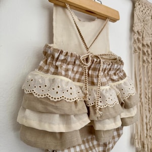 Puede incluir: Un pelele a cuadros beige y blanco con volantes en capas y ribete de encaje calado. El pelele está colgado de una percha de madera con una atadura de cuerda natural. El estilo general es bohemio y vintage.