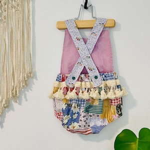 May include: A pink and white patchwork baby romper with a ruffled hem and tassel trim. The romper has a criss-cross strap design in the back.