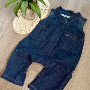 May include: A sleeveless denim jumpsuit in a medium wash. The jumpsuit features a button-up front, two chest pockets with zippers, and two hip pockets. The garment has a relaxed fit and is suitable for casual wear.