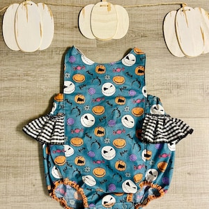 May include: A teal blue baby onesie with a Halloween theme. The onesie has a pattern of Jack Skellington's face, pumpkins, bats, and candy. The sleeves have black and white checkered ruffles.