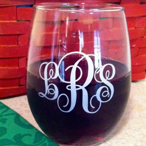 May include: A clear glass wine glass with a white monogram of the letters "B R S" etched on the side. The glass is filled with red wine and is sitting on a green and white patterned surface.