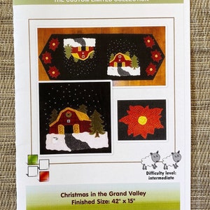 May include: A Laura's Home sewing pattern booklet titled Christmas in the Grand Valley. The cover features a black table runner with red barns, green pine trees, and red poinsettias. It includes a finished size of 42 inches by 15 inches.