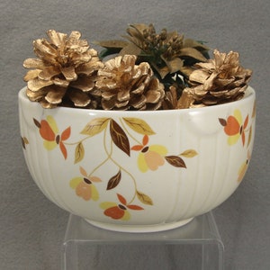 May include: A cream-colored ceramic bowl with vertical ribbing and a floral pattern in shades of orange, yellow, and brown. The bowl is filled with several metallic gold-painted pinecones and a gold-toned artificial poinsettia flower.