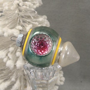 May include: Vintage Christmas ornament with a green and silver body, yellow stripes, and a red center. The ornament is displayed on a clear glass stand, with a white Christmas tree in the background.