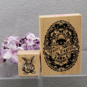 May include: Two wooden Easter stamps. One rectangular stamp features an ornate oval design with two rabbits, a basket, and the words "Happy Easter." The other stamp is square and shows a rabbit holding an Easter egg. Purple flowers are in the background.
