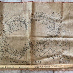 May include: A tan burlap rug hooking pattern featuring a hand-drawn design of two mirrored leafy fern branches. The text "NOVA SCOTIA MAT" and "19 X 31 1/2" is printed at the bottom. A wooden ruler is placed along the bottom edge for scale.
