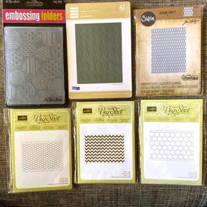 May include: A collection of embossing folders in various designs, including circles, diamonds, and geometric patterns. The folders are in clear plastic packaging, with some featuring the "Big Shot" and "Sizzix" brands. The color palette includes clear, gold, and brown.