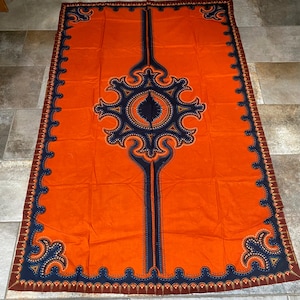 May include: An orange textile with a large, symmetrical design in navy blue and brown. The central motif is a stylized floral pattern, with a vertical navy blue stripe down the center. The edges are trimmed with a decorative border.