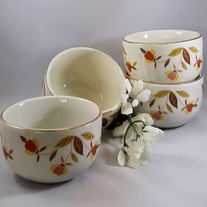 May include: A set of three off-white ceramic bowls featuring a delicate floral pattern in shades of orange, yellow, and brown. Each bowl has a thin gold-toned rim. A small cluster of white artificial flowers is placed between the bowls.