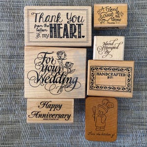 May include: A collection of six rectangular wooden rubber stamps featuring various black ink designs and text including Thank You from the bottom of my HEART, For your Wedding with roses, Happy Anniversary, A Friend loves at all times, Hand Stamped By, and Handcrafted By with a chef bear illustration.