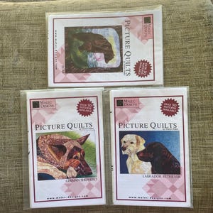 May include: Three rectangular quilt pattern packets by Malec Designs titled Picture Quilts. The top packet features a bear, the bottom left a German Shepherd, and the bottom right a Labrador Retriever. Each white packet has pink accents.