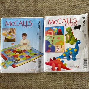 May include: Two McCall's Crafts sewing pattern envelopes, M7104 and M7553. M7104 features a colorful patchwork play mat with animal appliques. M7553 displays three plush dinosaur toys in green, blue with yellow spots, and red, alongside a landscape quilt.