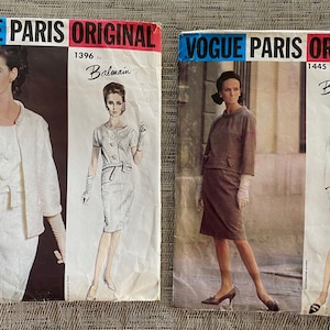 May include: Two vintage Vogue Paris Original sewing pattern envelopes. One features a white suit with a jacket and dress, the other a brown skirt suit. Both are by Balmain, with the Vogue Paris Original logo at the top.