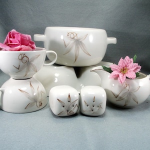 May include: A white ceramic dinnerware set featuring a brown floral pattern with curly stems. The set includes a lidded tureen, two cups, a creamer, and salt and pepper shakers. Pink artificial flowers are placed inside one cup and the creamer.