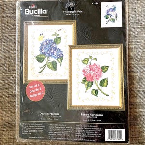 May include: A Plaid Bucilla crewel embroidery kit titled Hydrangea Pair, featuring two framed designs of blue and pink hydrangea flowers with green leaves. The package includes a set of two 6.25 inch by 8.25 inch stamped fabric pieces.