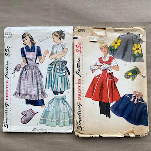 May include: Two vintage Simplicity sewing pattern envelopes, numbers 2295 and 4899. The left envelope features illustrations of aprons and oven mitts. The right envelope displays a red apron, a gray skirt with yellow flowers, and a blue apron.