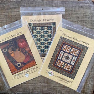 May include: Three Thimbleberries quilt pattern packets in plastic sleeves. The packets are titled "Autumn Medley", "Cottage Flower", and "Prairie Blossoms". Each packet features a quilt design with various colors and patterns. The brand name "Thimbleberries" is visible on each packet.