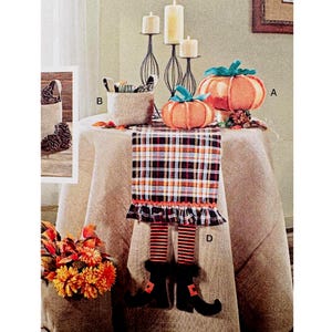 May include: A festive Halloween table setting featuring orange fabric pumpkins with green accents, a plaid table runner with witch legs, and tall black candle holders. The table is set for autumn with fall foliage.