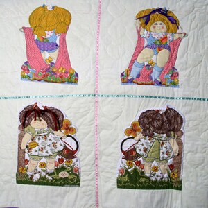 May include: A handmade quilt featuring four appliqued doll figures. The dolls have yellow or brown hair, wearing dresses and holding accessories. The quilt is white with pink and green accents, and the dolls are surrounded by floral designs.