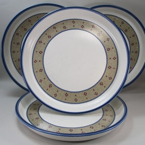 May include: Four white ceramic dinner plates with a blue rim and a decorative band of tan and red geometric patterns. The plates are stacked and arranged to show the design.