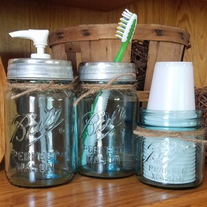 May include: A set of three blue mason jars with silver lids, twine accents, and the text "Ball Perfect Mason." One jar has a white soap dispenser, another holds a toothbrush, and the third contains white cups. A rustic bathroom organizer.