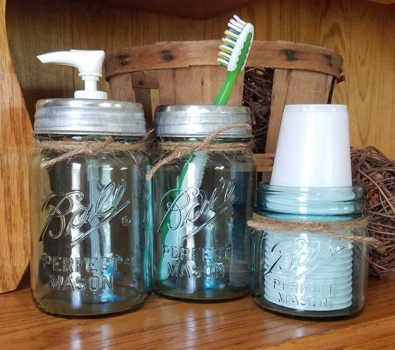 May include: A set of three blue mason jars with silver lids, twine accents, and the text "Ball Perfect Mason." One jar has a white soap dispenser, another holds a toothbrush, and the third contains white cups. A rustic bathroom organizer.