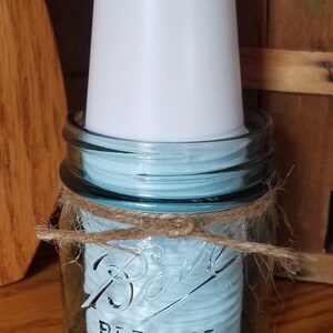 May include: A clear glass Ball Perfect Mason jar with a light blue interior, tied with twine. A white plastic cup is placed inside the jar. The jar is sitting on a wooden surface.