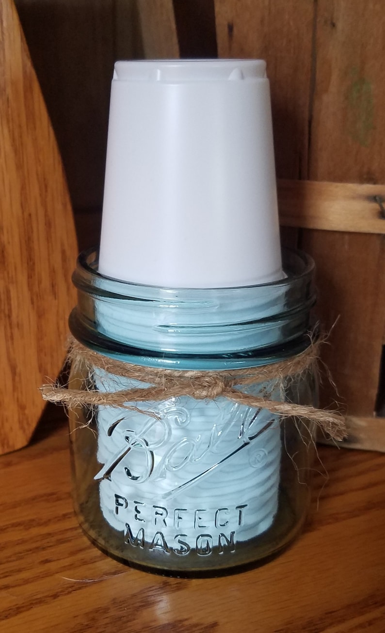 May include: A clear glass Ball Perfect Mason jar with a light blue interior, tied with twine. A white plastic cup is placed inside the jar. The jar is sitting on a wooden surface.